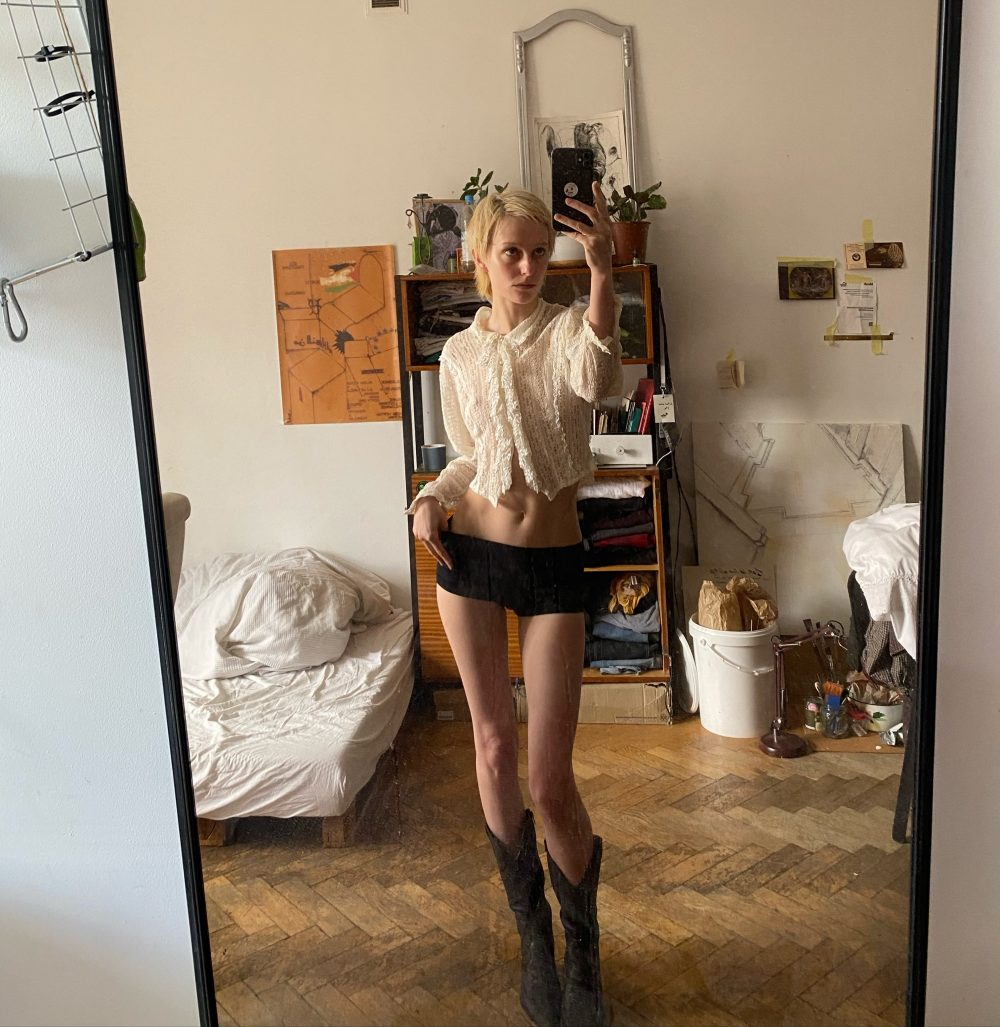 Person taking a mirror selfie in a cluttered room, wearing a white lace blouse and black shorts with tall black boots.
