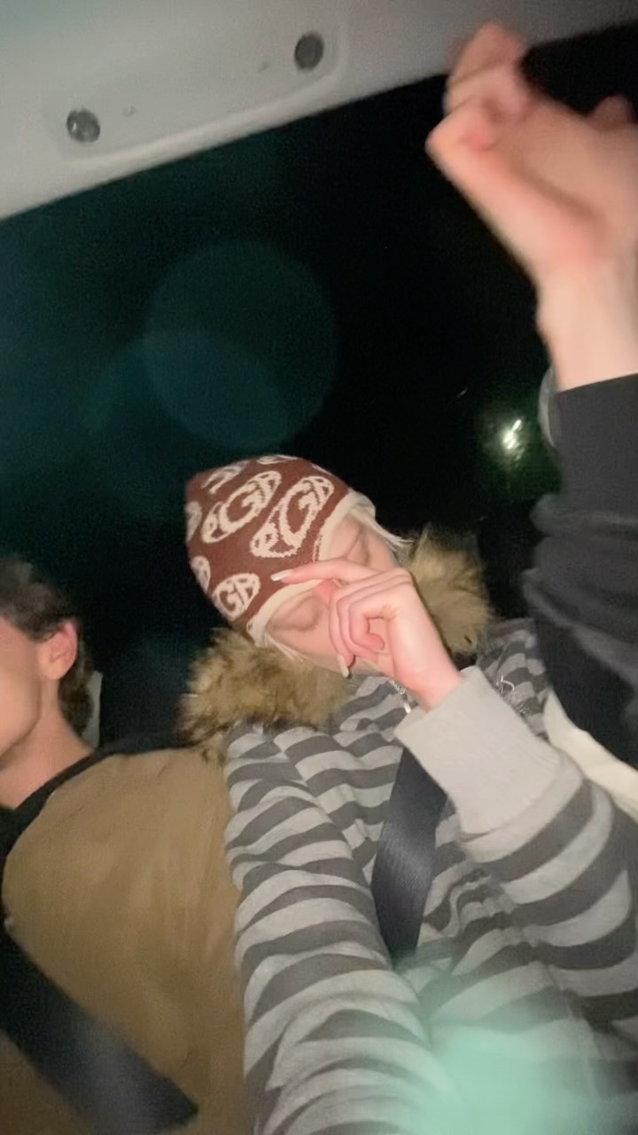 Two people ride in a dark car at night; one in a striped hoodie and brown patterned beanie making a peace sign near their face, with a seatbelt visible, another person partially seen beside them.