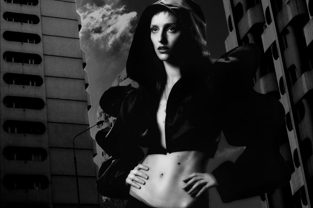 Monochrome mural of a woman with a hooded cloak, hands on hips, painted on a large wall between city buildings.