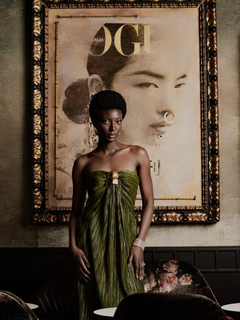 Elegant Black woman with a natural hairstyle wearing a green strapless dress, standing in front of a large ornate framed portrait/artwork.