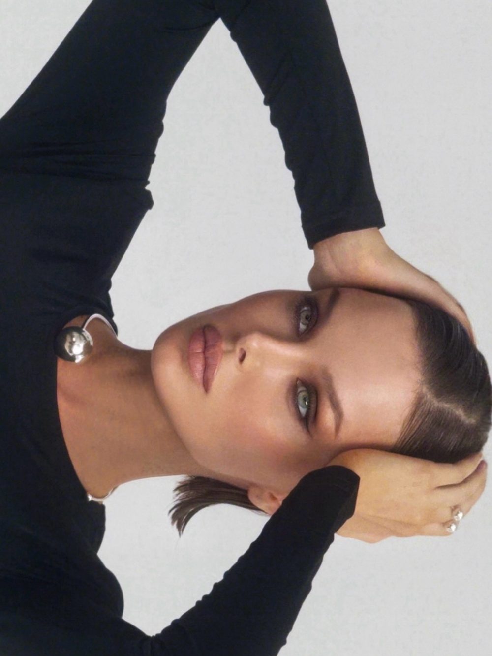 Portrait of a woman lying on her side with arms raised, wearing a black long-sleeve top and a silver necklace, looking at the camera.