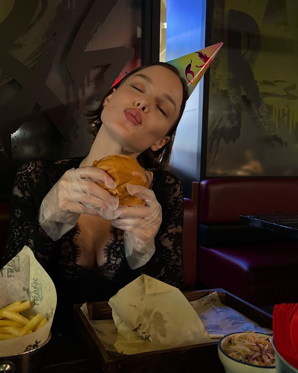 Woman wearing a rainbow party hat puckers her lips as she rests a sandwich against her cheek, with fries on the table nearby.