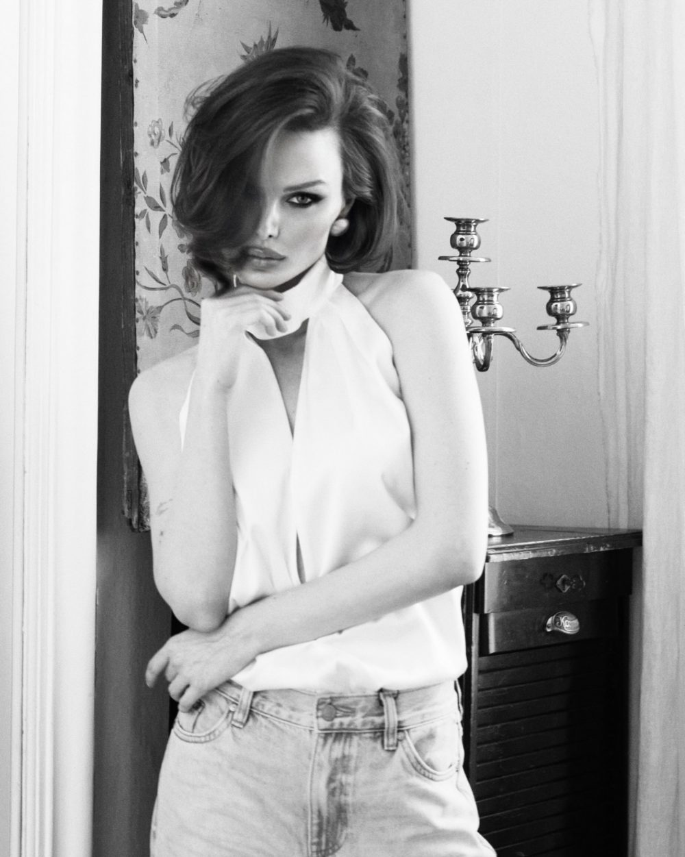 Woman in a white halter top stands beside a vintage candelabra on a dresser, posed near a decorative wall panel in a black-and-white photo