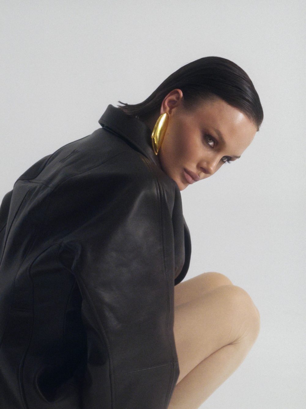 Side profile of a woman wearing a black leather jacket with a large gold hoop earring, against a pale background.