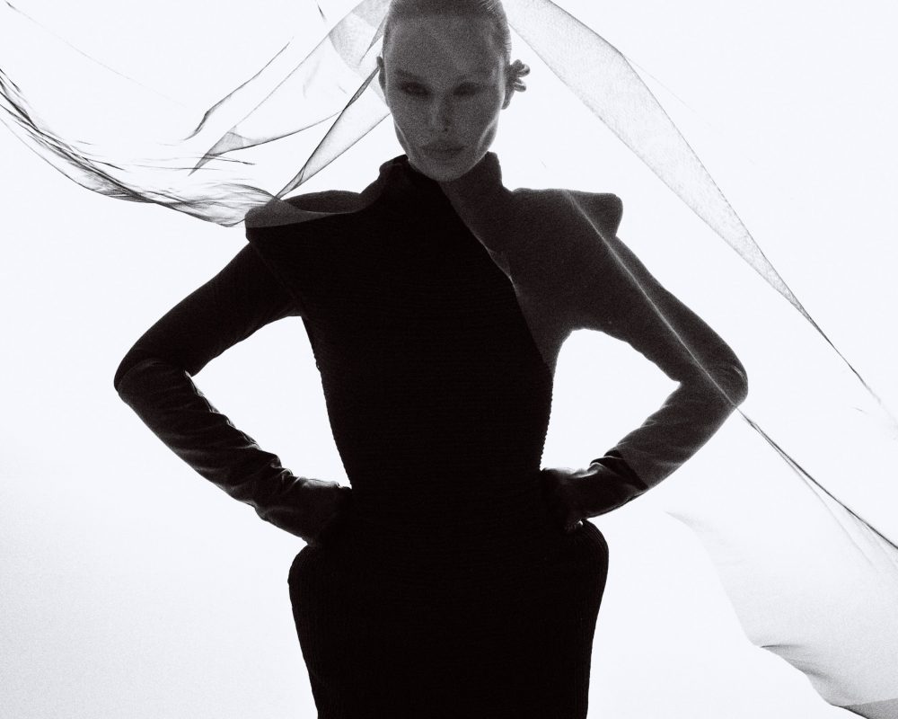 Silhouette of a person in a black dress with hands on hips and fabric swirling around the head in a high-contrast image.