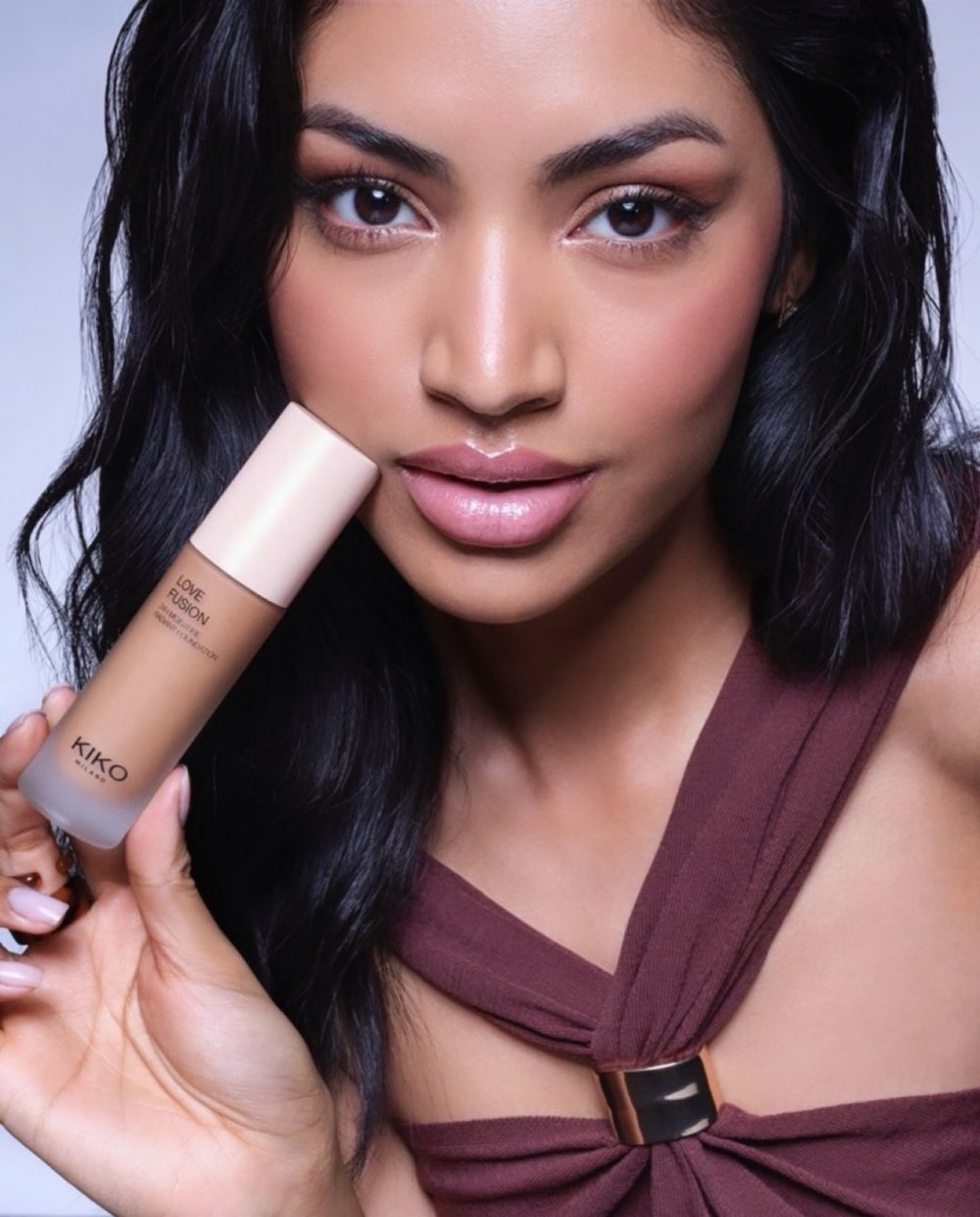 Close-up portrait of a woman with polished makeup holding a KIKO Milano Love Fusion foundation bottle near her cheek.