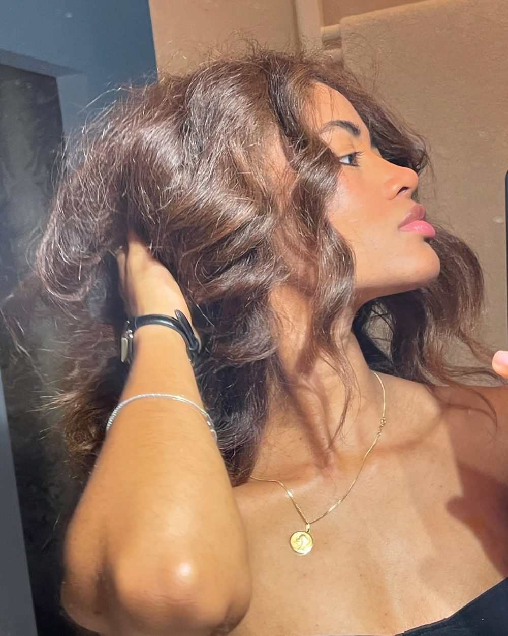 Young woman with wavy brown hair seen in profile, lifting her hair back with both hands, wearing a gold necklace and a black top in a bright setting.