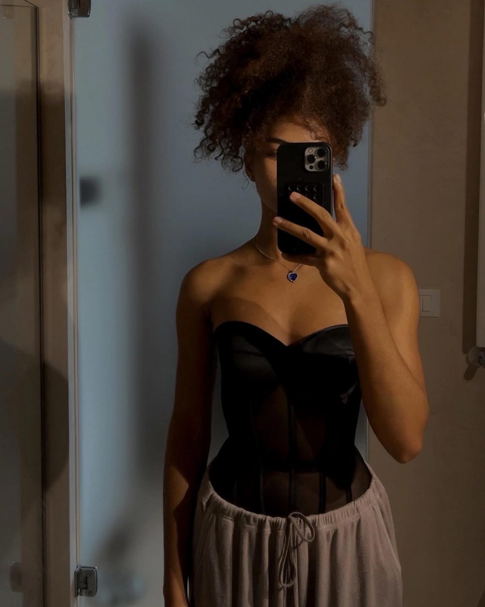 Person taking a mirror selfie in a bathroom, wearing a black strapless corset top and light gray drawstring pants, with a blue heart pendant necklace and natural curly hair.