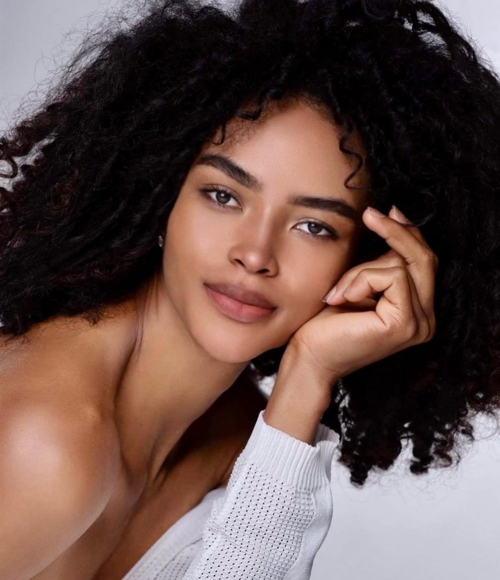 Close-up portrait of a woman with dark, voluminous curly hair resting her chin on her hand.