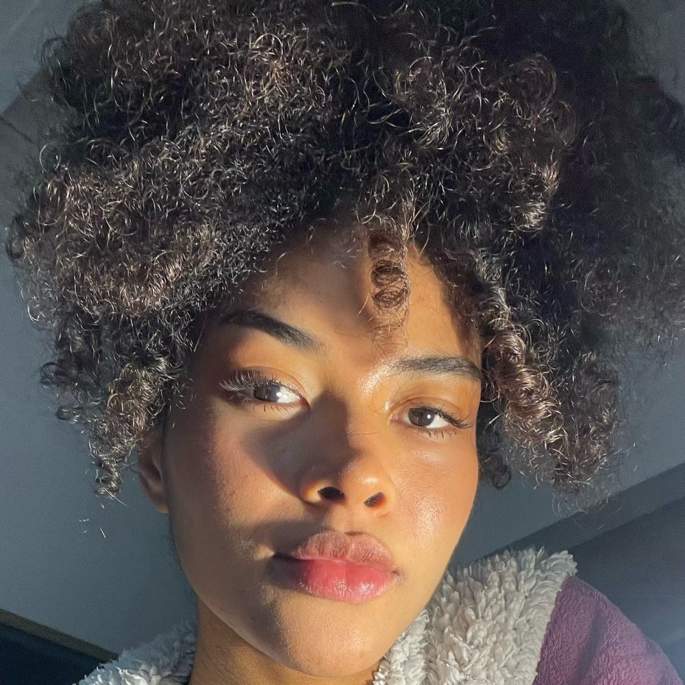 Close-up portrait of a person with dark skin and tight curly hair illuminated by warm sunlight, wearing a purple garment with a white fleece collar.