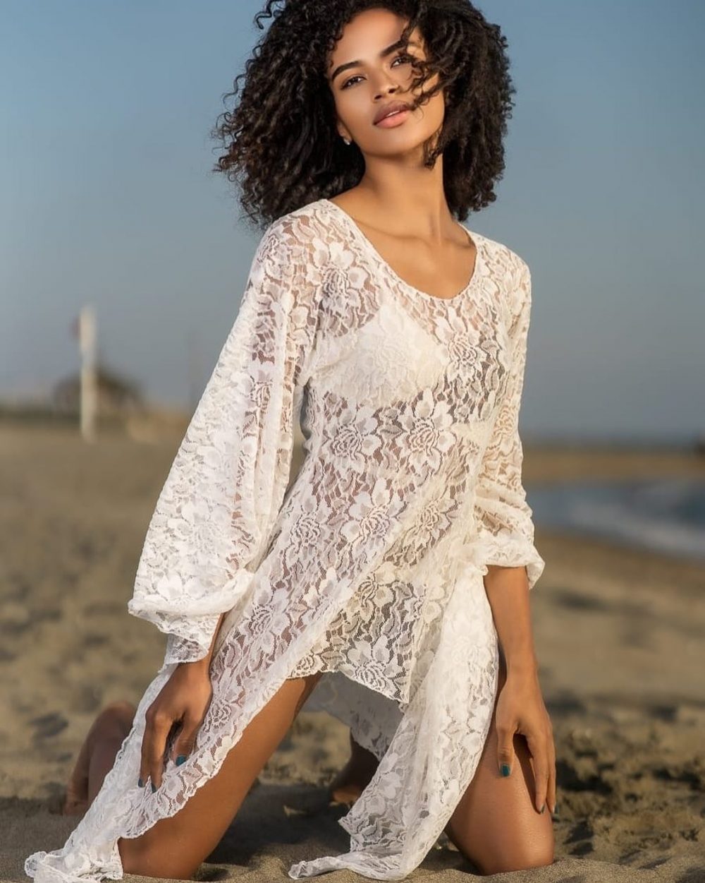 Woman posing on a sandy beach in a white lace dress, hair blown by the wind, looking toward the camera.
