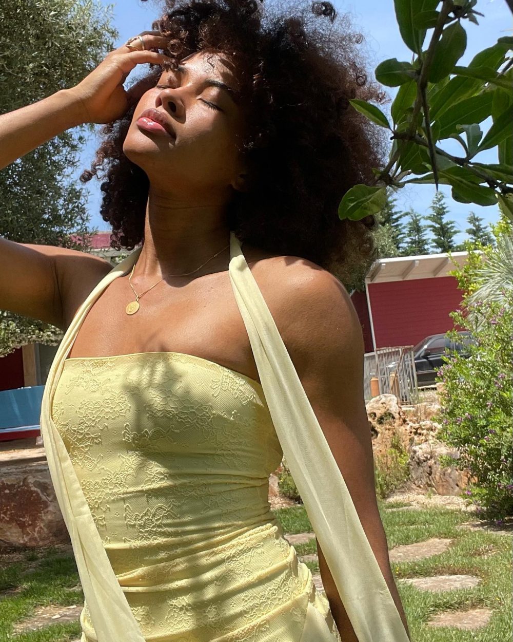 Woman in a pale yellow lace dress stands outdoors, eyes closed, hand on her forehead, sunlit garden background.