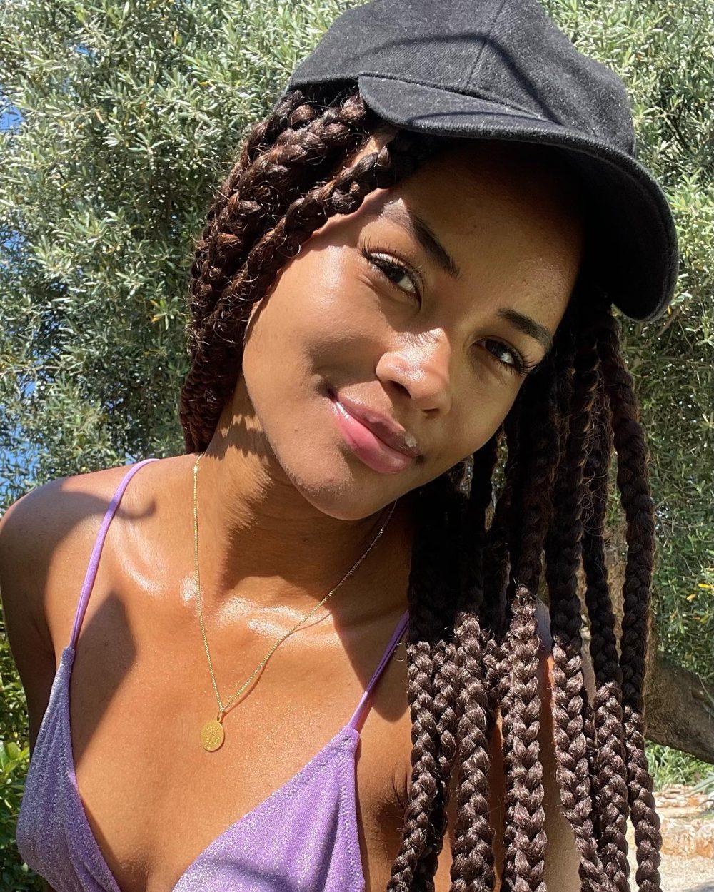 Close-up selfie of a smiling woman with long braided hair wearing a black cap outdoors on a sunny day.
