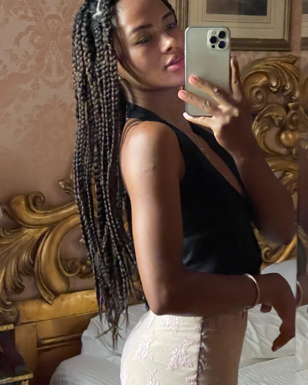 Woman with long braids wearing a black sleeveless top and white lace shorts, taking a mirror selfie in a bedroom with an ornate headboard.