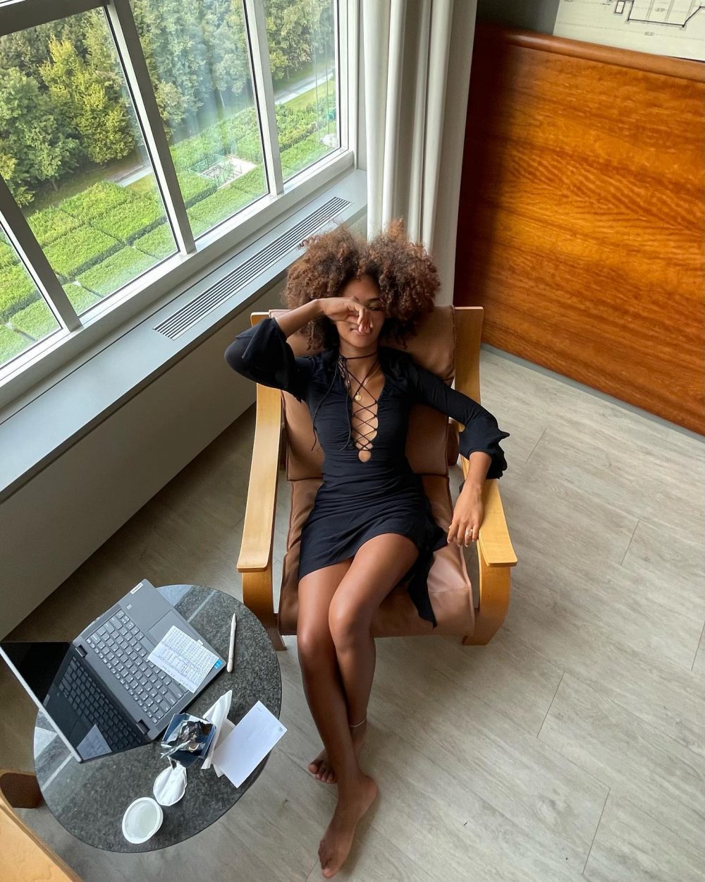 Woman with curly hair in a black lace-up dress relaxing in a wooden lounge chair by a large window, covering her eyes with one hand.