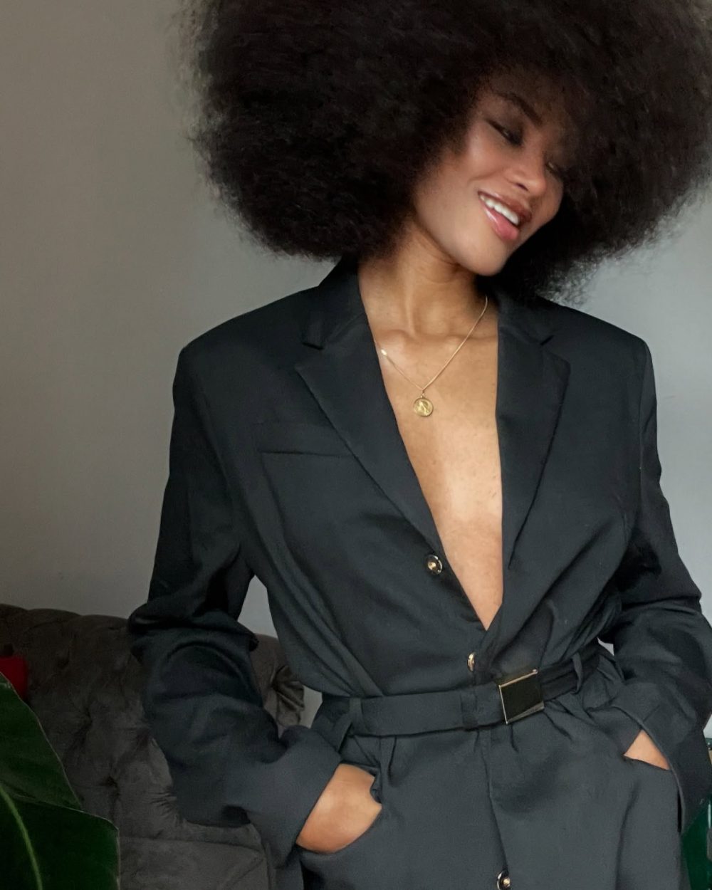 Woman with a large natural afro wearing a black blazer and gold pendant necklace, smiling with head tilted to the side.