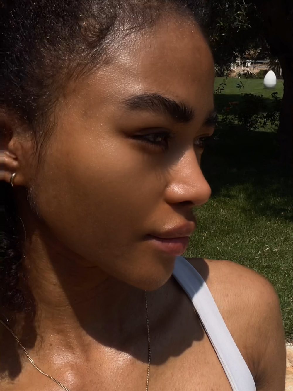 Side profile of a woman outdoors in bright sunlight, wearing a white tank top and a thin necklace, with a grassy park and a white egg-shaped sculpture in the background.