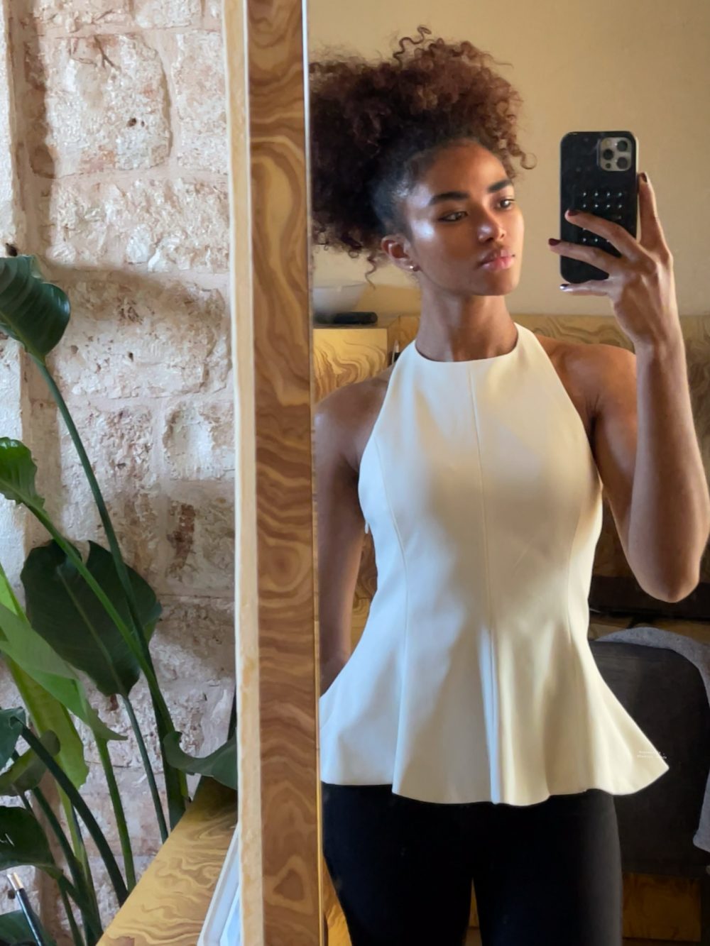 Woman taking a mirror selfie in a white halter peplum top and black pants, with a plant and stone wall in the background.