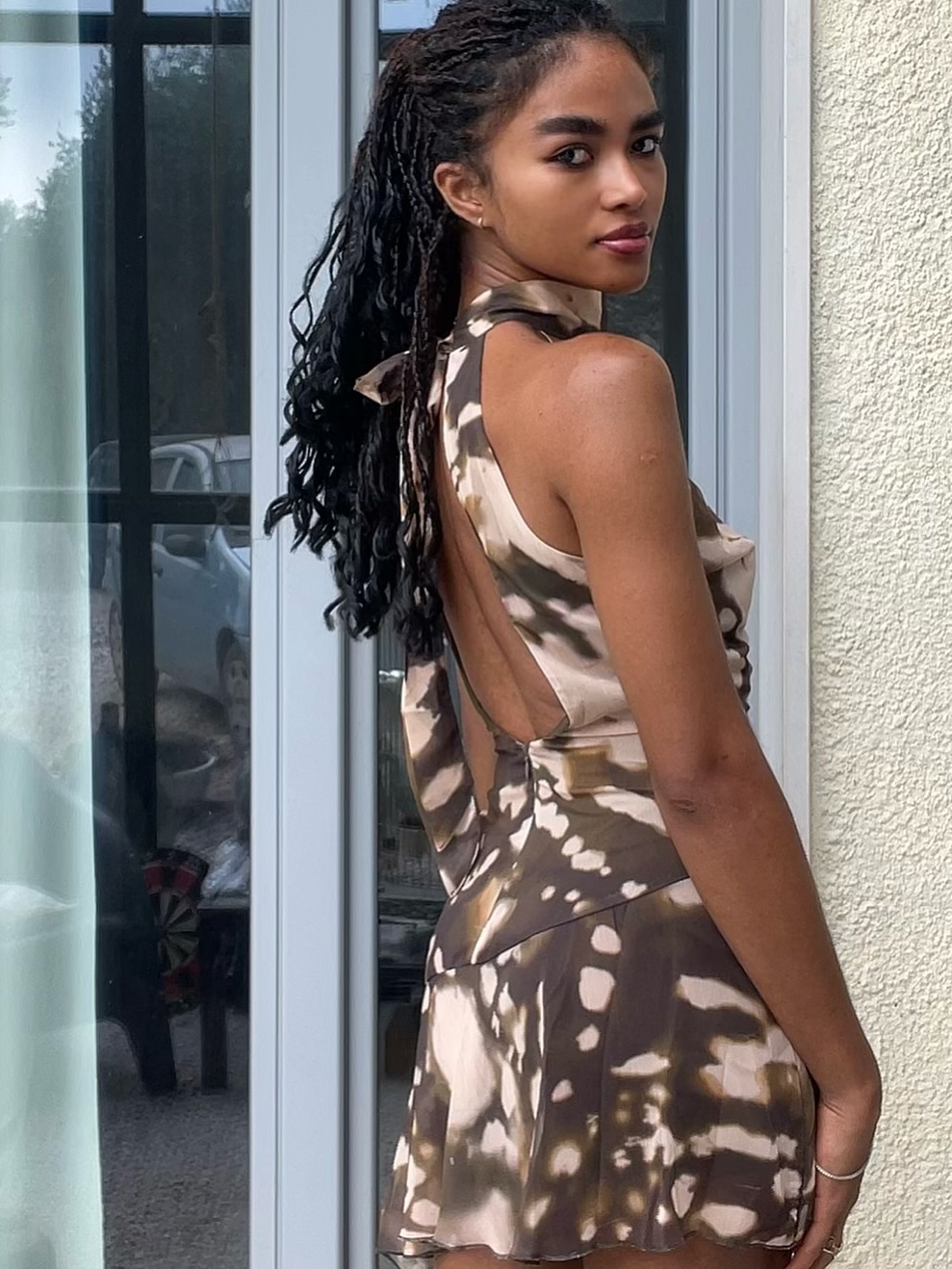 Woman in a sleeveless halter-back tie-dye dress standing outdoors beside a glass door, glancing over her shoulder.