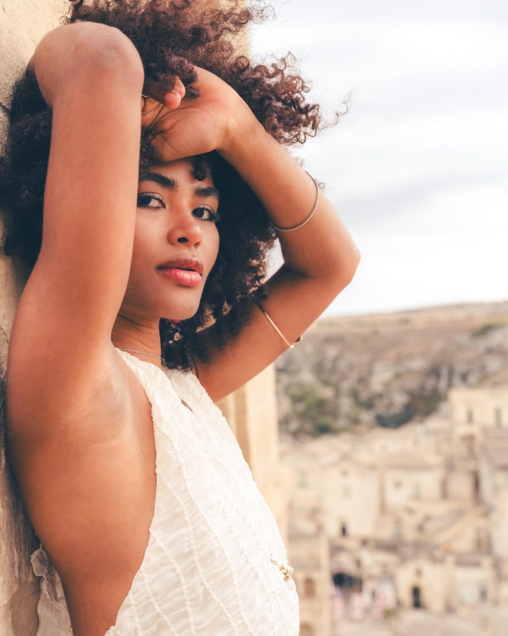 Young woman with curly hair leaning against a stone wall, arms raised above her head, wearing a white textured top, with a historic cityscape in the blurred background.