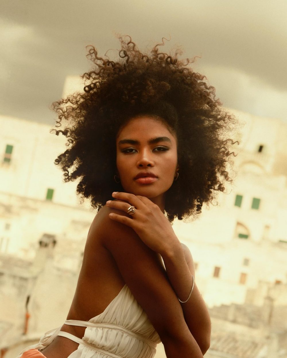 Portrait of a woman with large curly hair wearing a light wrap dress, posing outdoors against a pale urban backdrop.