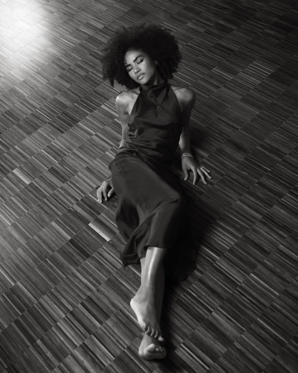 Black-and-white portrait of a woman in a satin halter dress reclining on a striped floor, eyes closed in a serene pose.