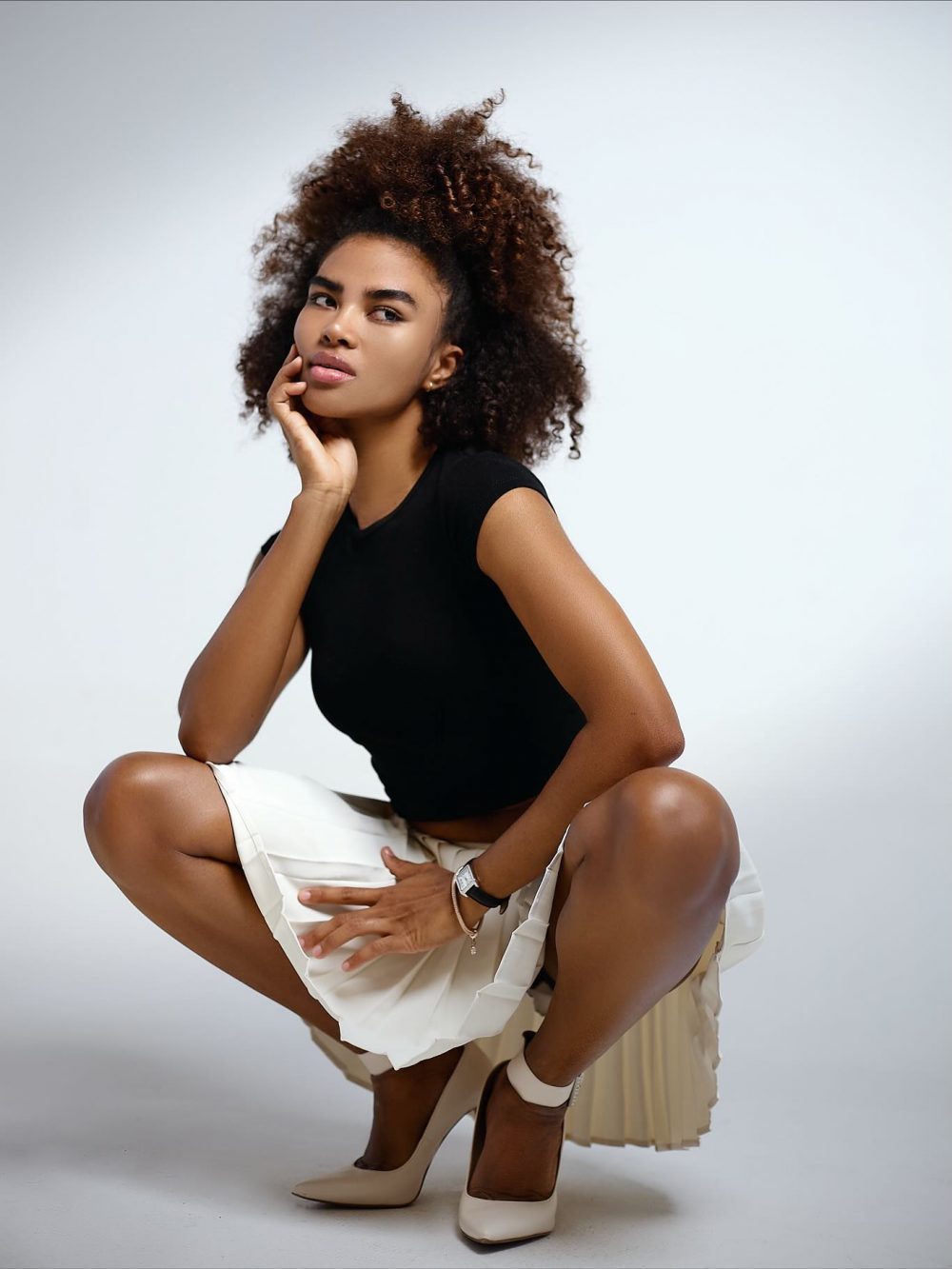 Woman posing in a black top, white pleated skirt, and nude heels, crouching with her hand resting on her chin against a light background.