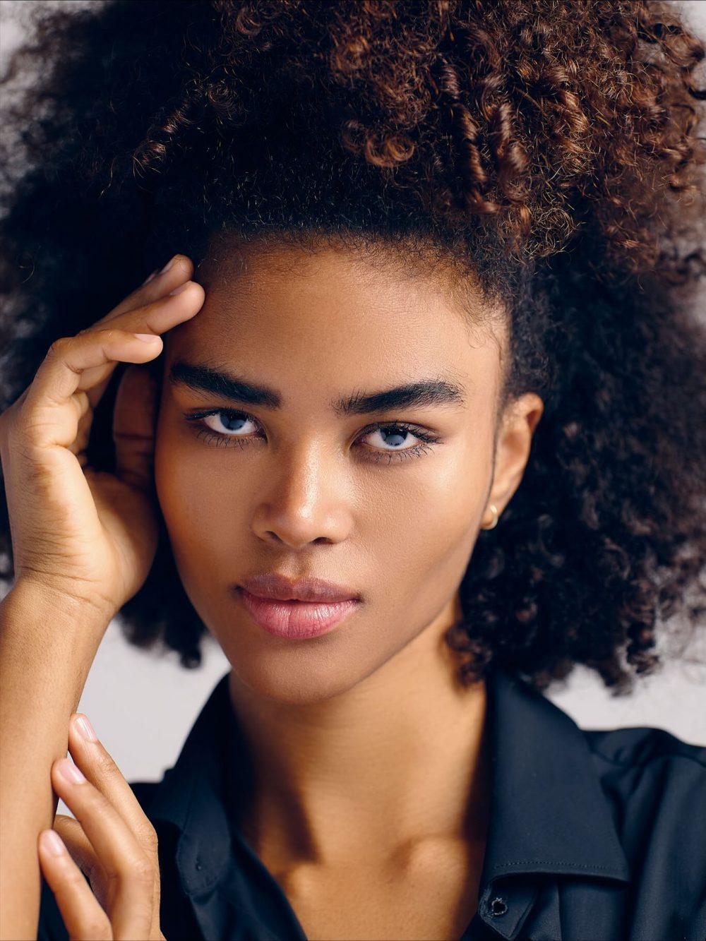 Close-up portrait of a woman with tight, curly hair resting her hand on the side of her head, looking directly at the camera.