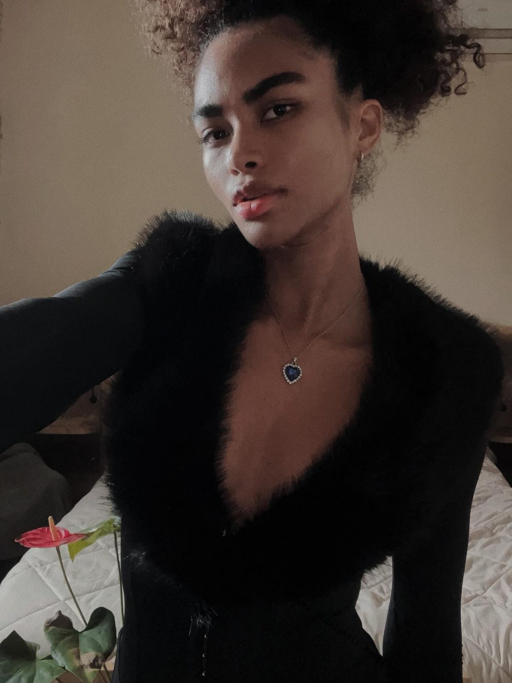 Person wearing a black furry outfit with a blue heart pendant necklace, posing in dim lighting.