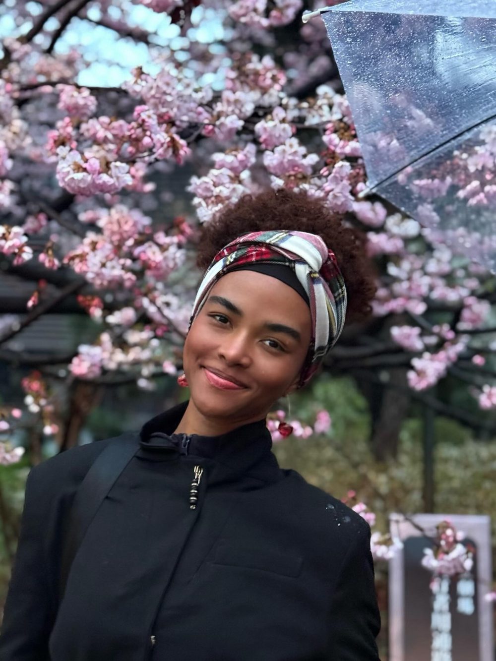 Portrait of a smiling person with a colorful headwrap among pink cherry blossoms and a raindrops-streaked umbrella nearby.