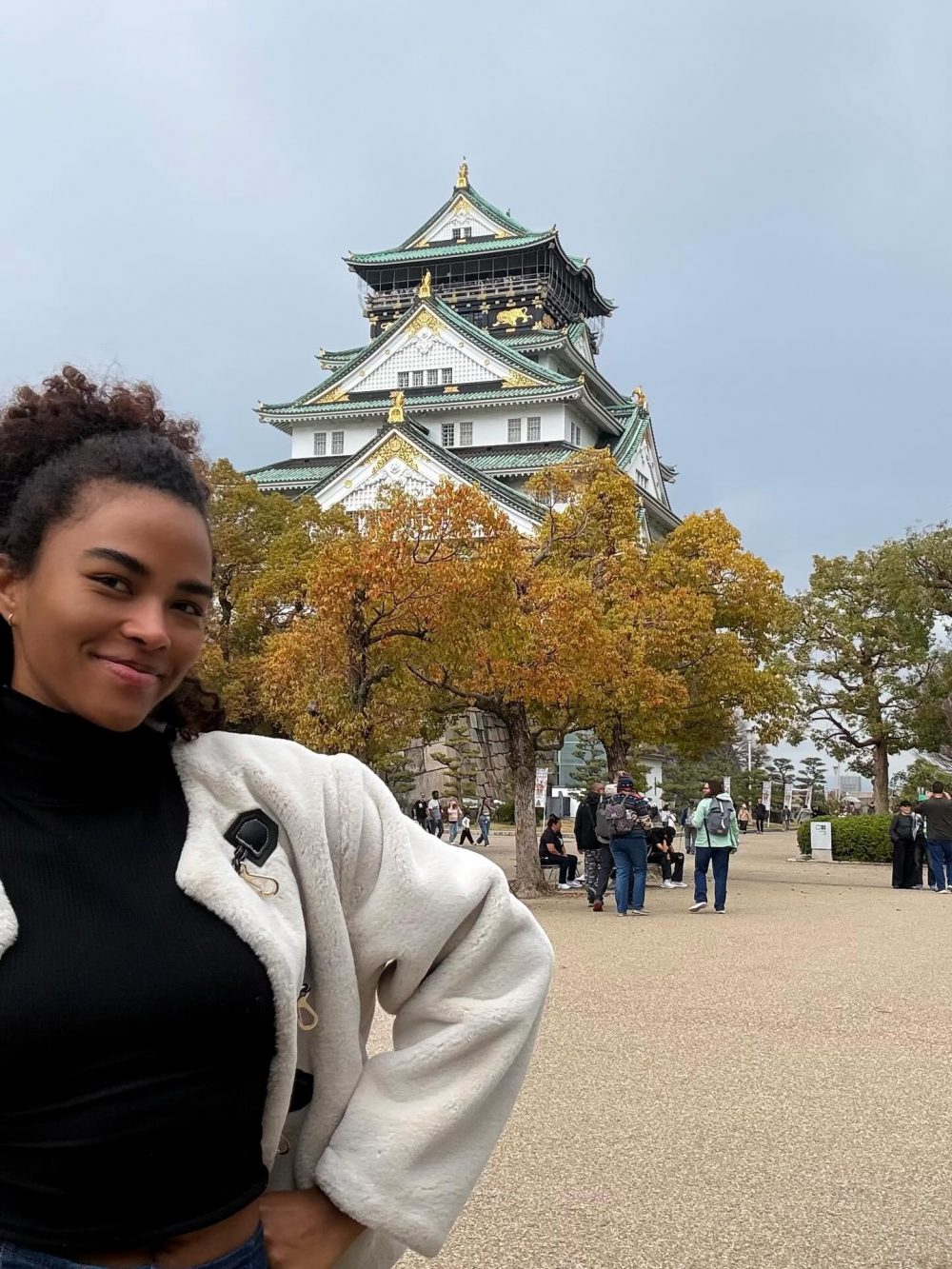 Young woman taking a selfie in front of a traditional Japanese castle with green tiled roofs and autumn trees in the foreground.