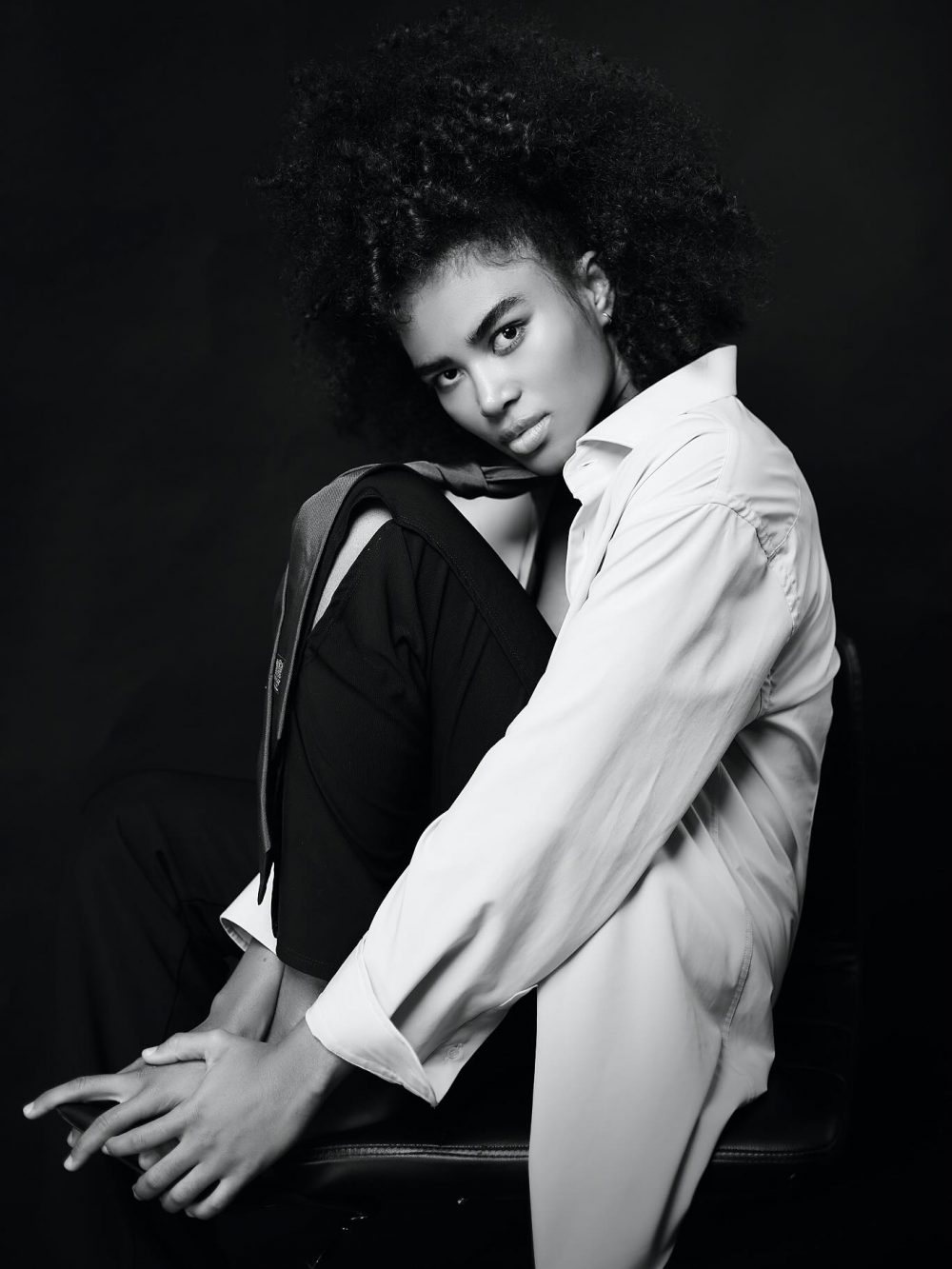 Black-and-white portrait of a woman with a large natural afro, wearing a white button-down shirt, seated sideways against a dark background and looking at the camera.