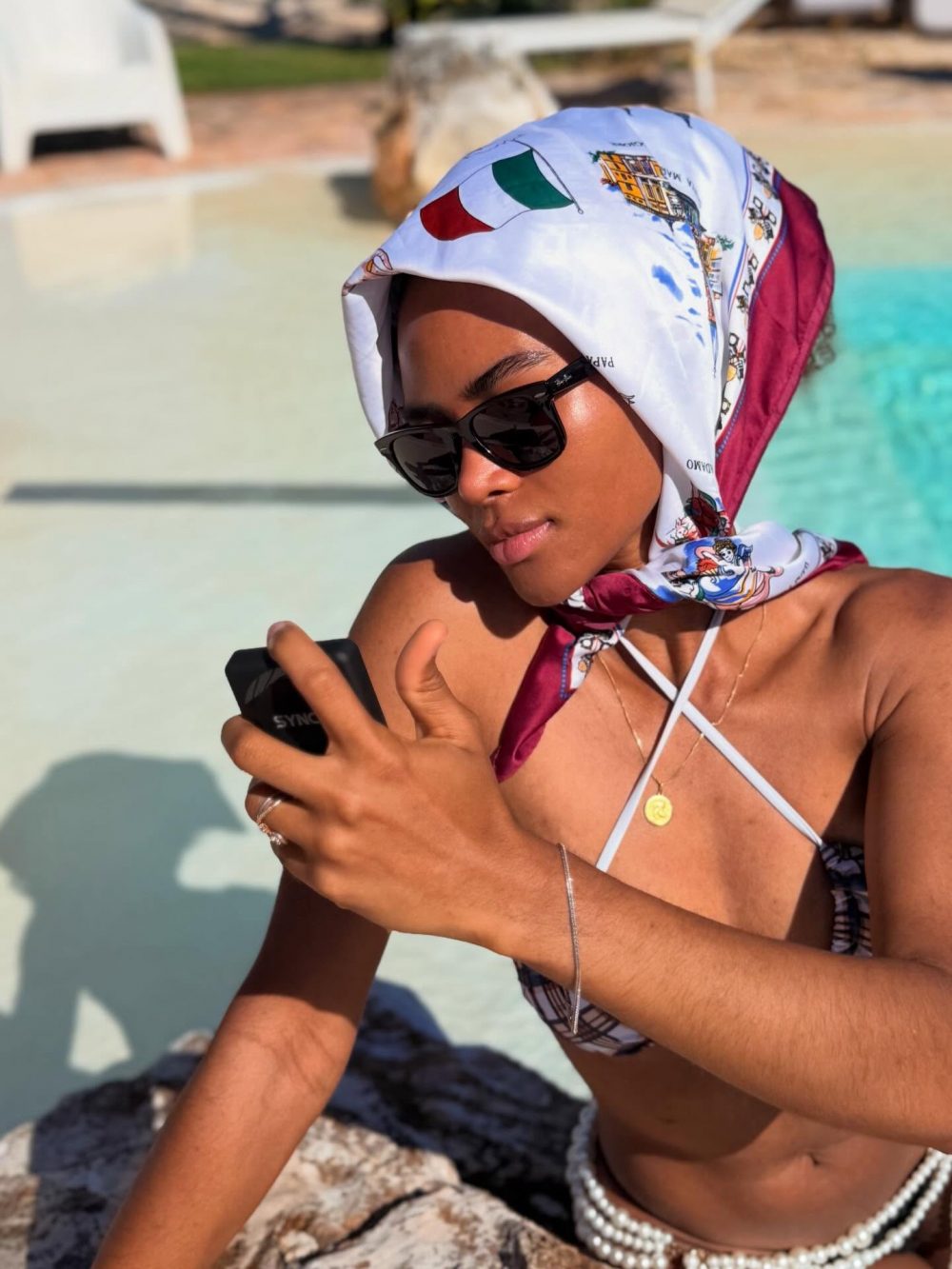 Woman wearing a patterned headscarf and black sunglasses by a pool, looking at a phone held in her hands.