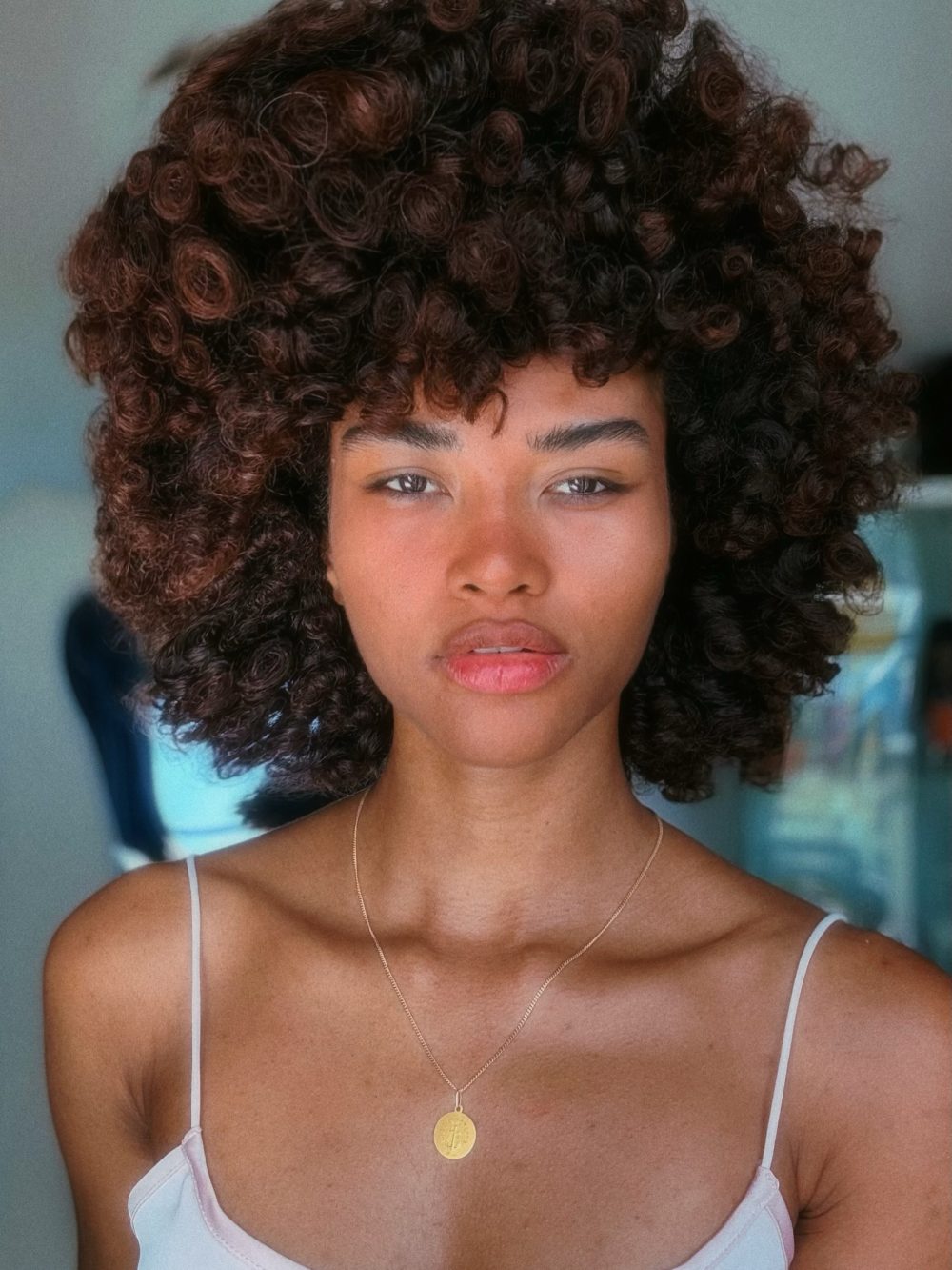 Portrait of a woman with a voluminous curly afro, wearing a white spaghetti-strap top and a gold pendant necklace.