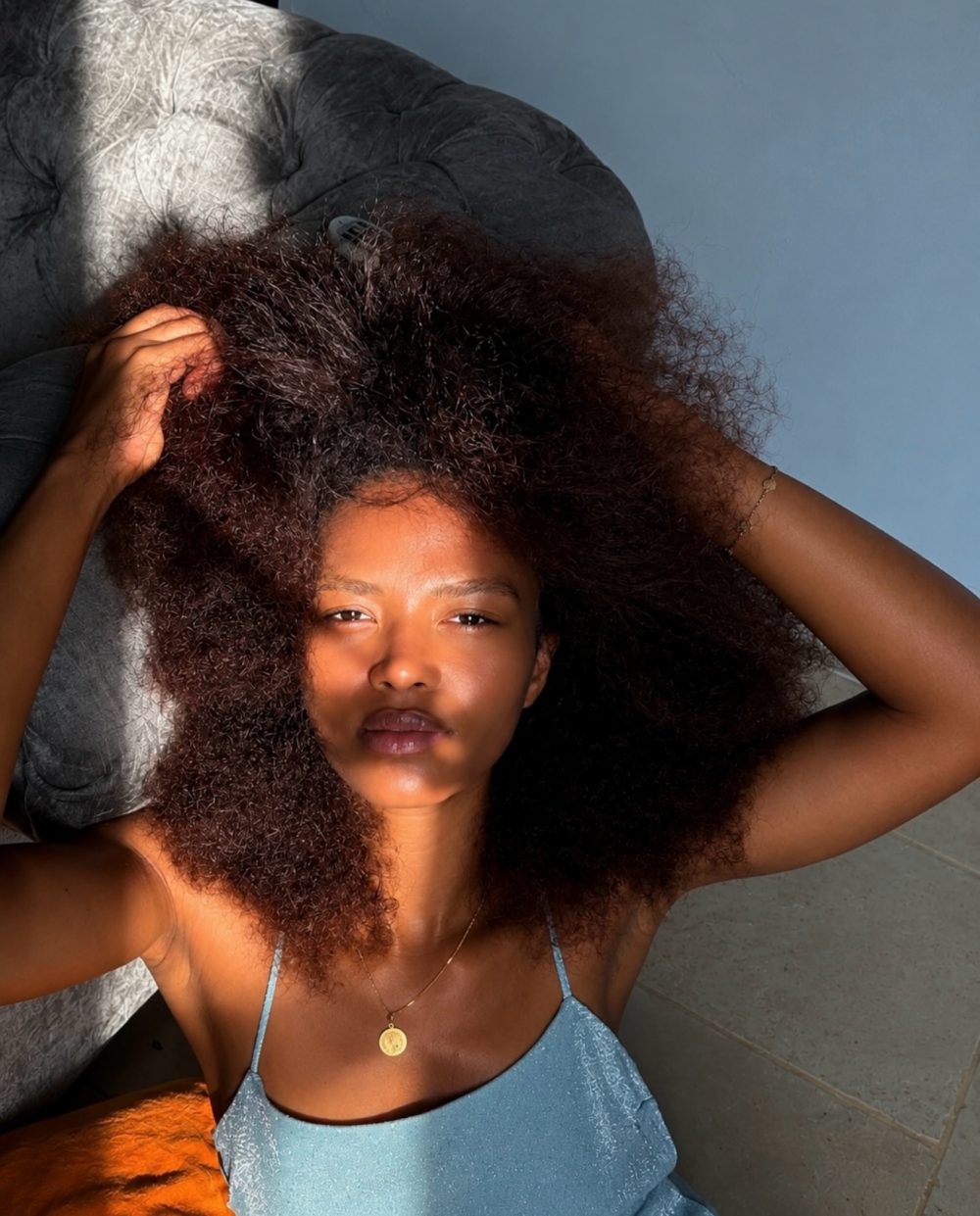 Young woman with a voluminous curly afro lying back, hands in her hair, wearing a blue spaghetti-strap top and a gold necklace.