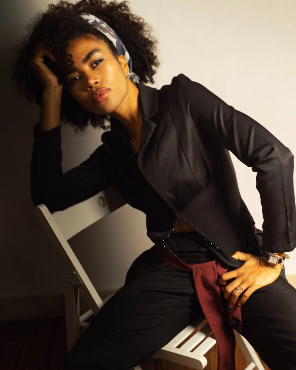 Portrait of a person in a black blazer and pants, leaning against a chair with one hand on their head, looking at the camera in a stylish studio shot.