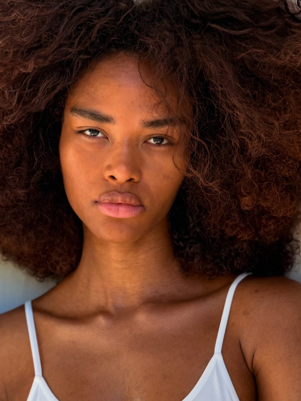 Close-up of a woman with voluminous curly hair surrounding her face, wearing a white spaghetti-strap top.