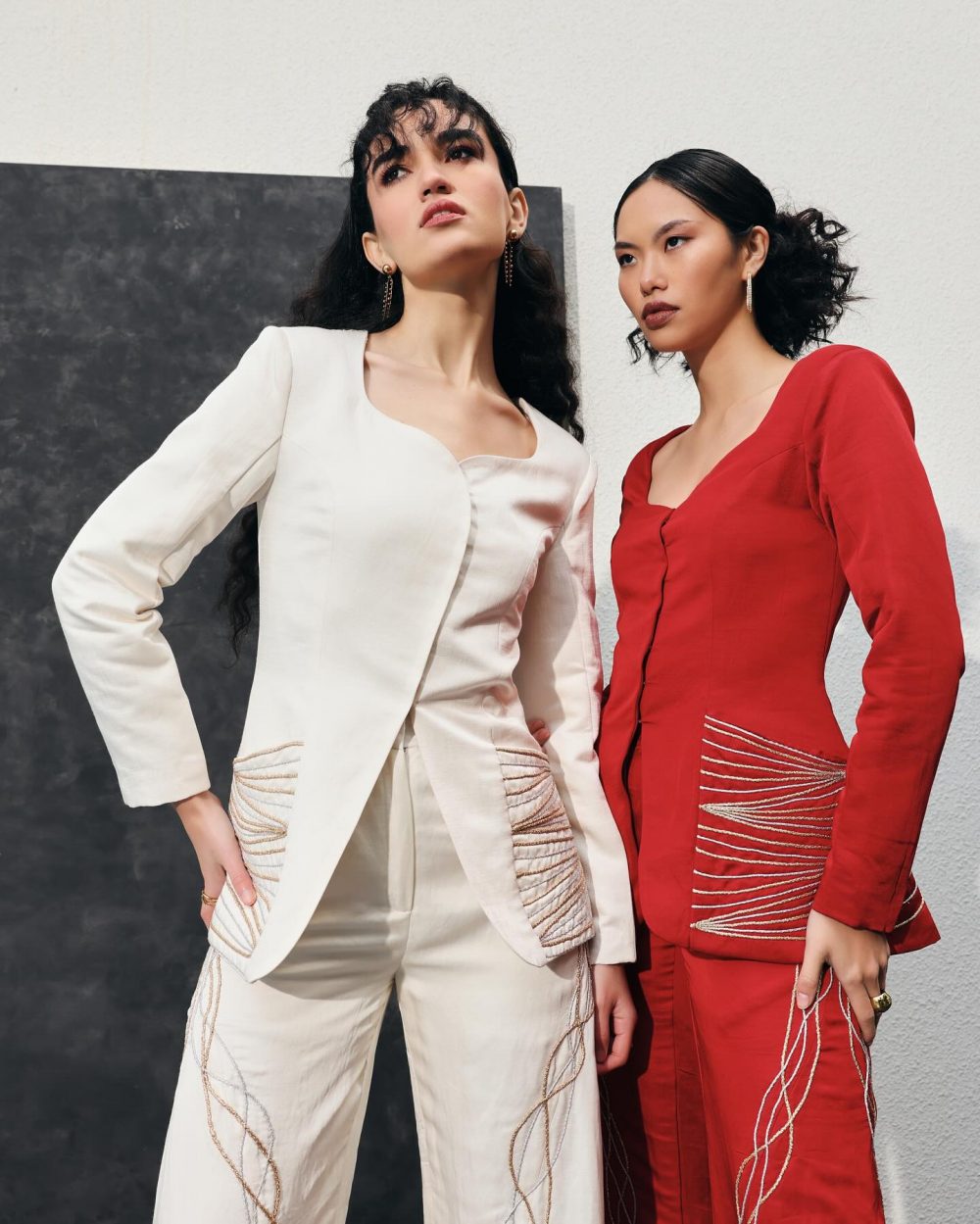 Two fashion models pose in cream and red tailored suits with gold embroidery against a light backdrop.