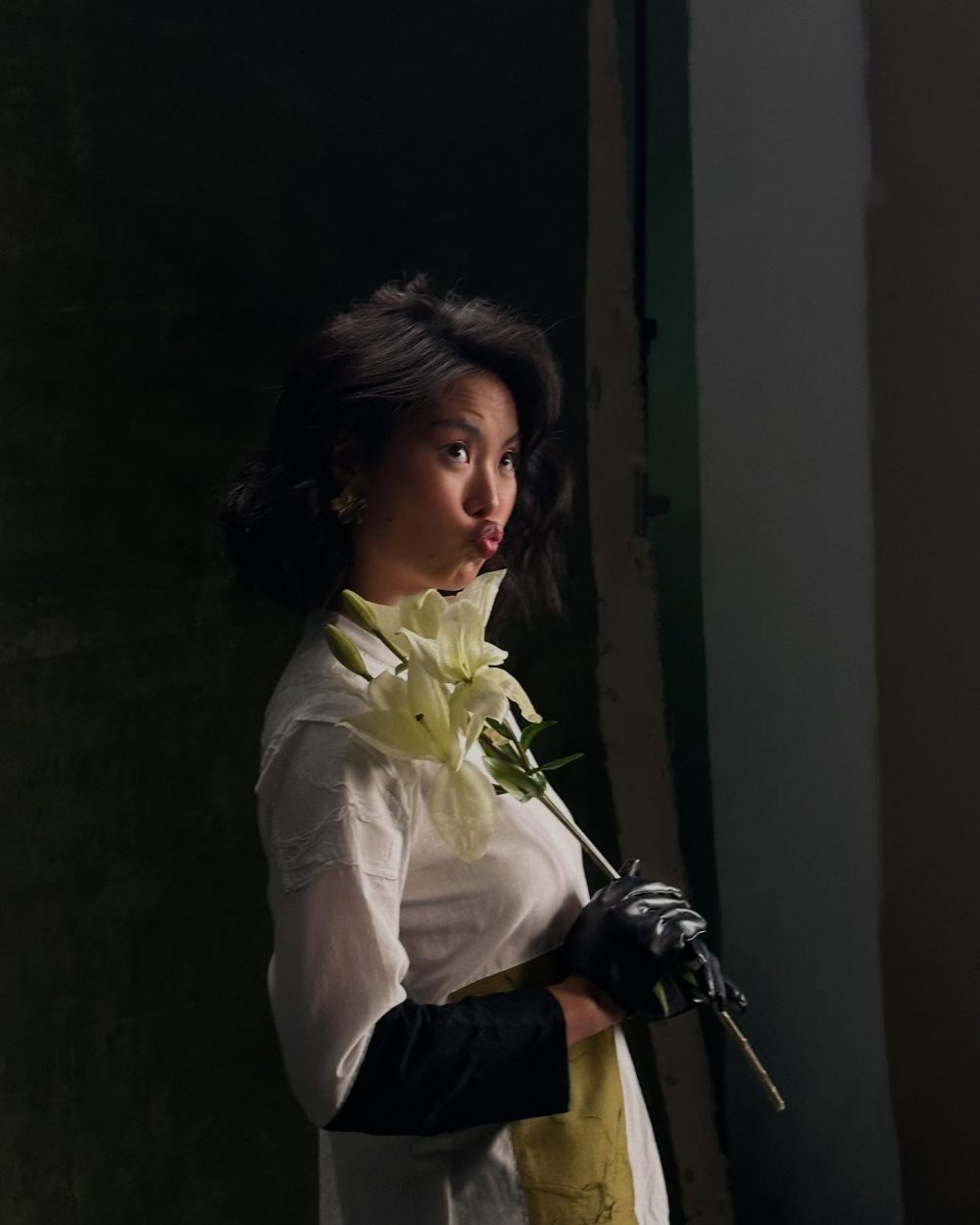Portrait of a woman in a light top and long black gloves, holding white lilies near her chest in a dark setting.