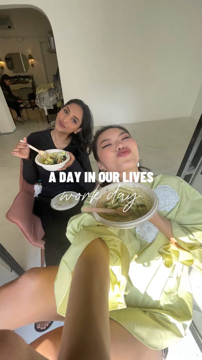Two women sit together, smiling and eating bowls of food, taking a selfie in a bright room during a work-day moment.