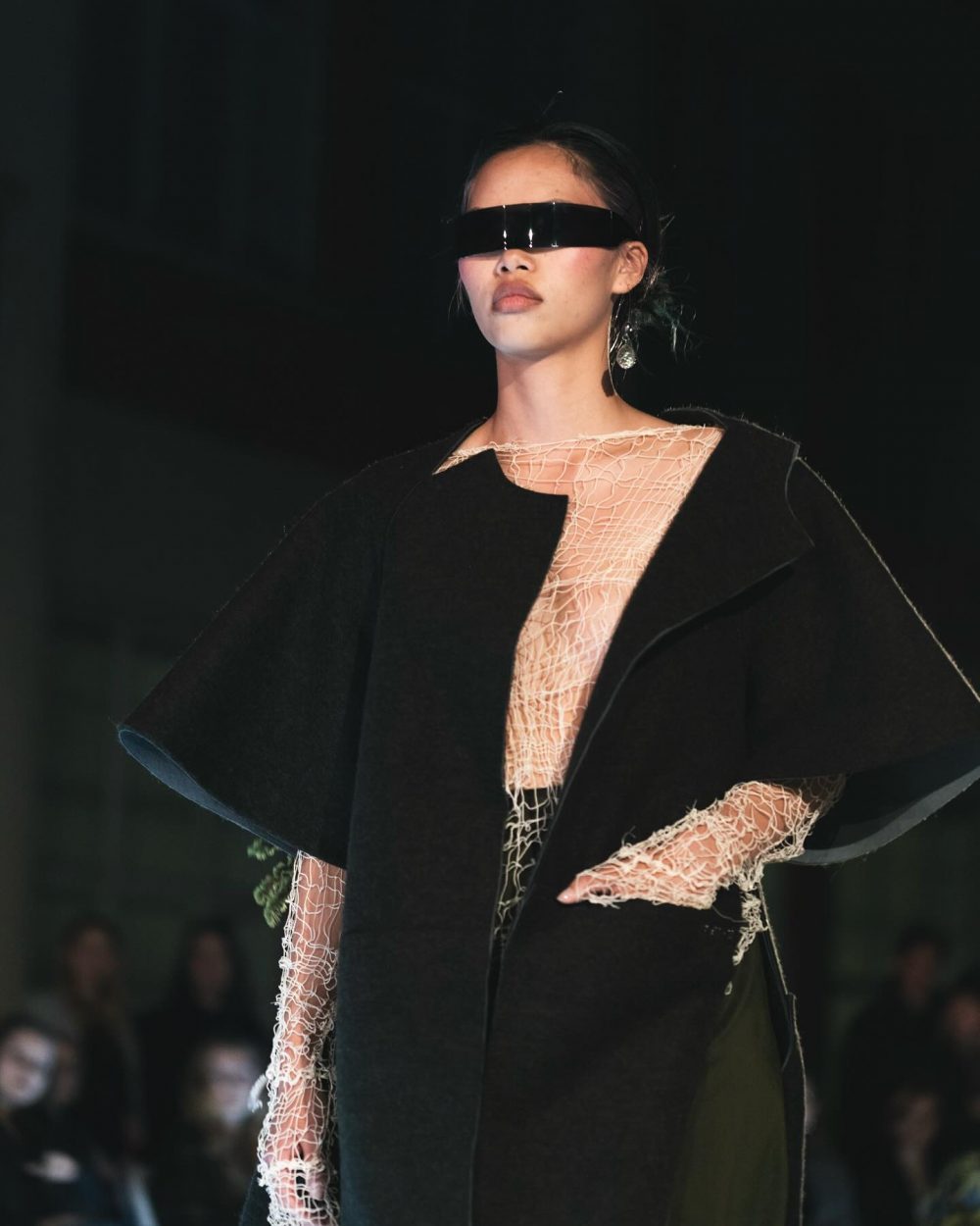 Model on a runway wearing an oversized black coat over a peach mesh top and a futuristic black eye visor that covers the eyes.