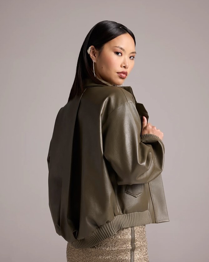 Woman in an olive-green leather bomber jacket, turned slightly away, looking over her shoulder at the camera. The jacket has a ribbed hem and a back ventilation seam.