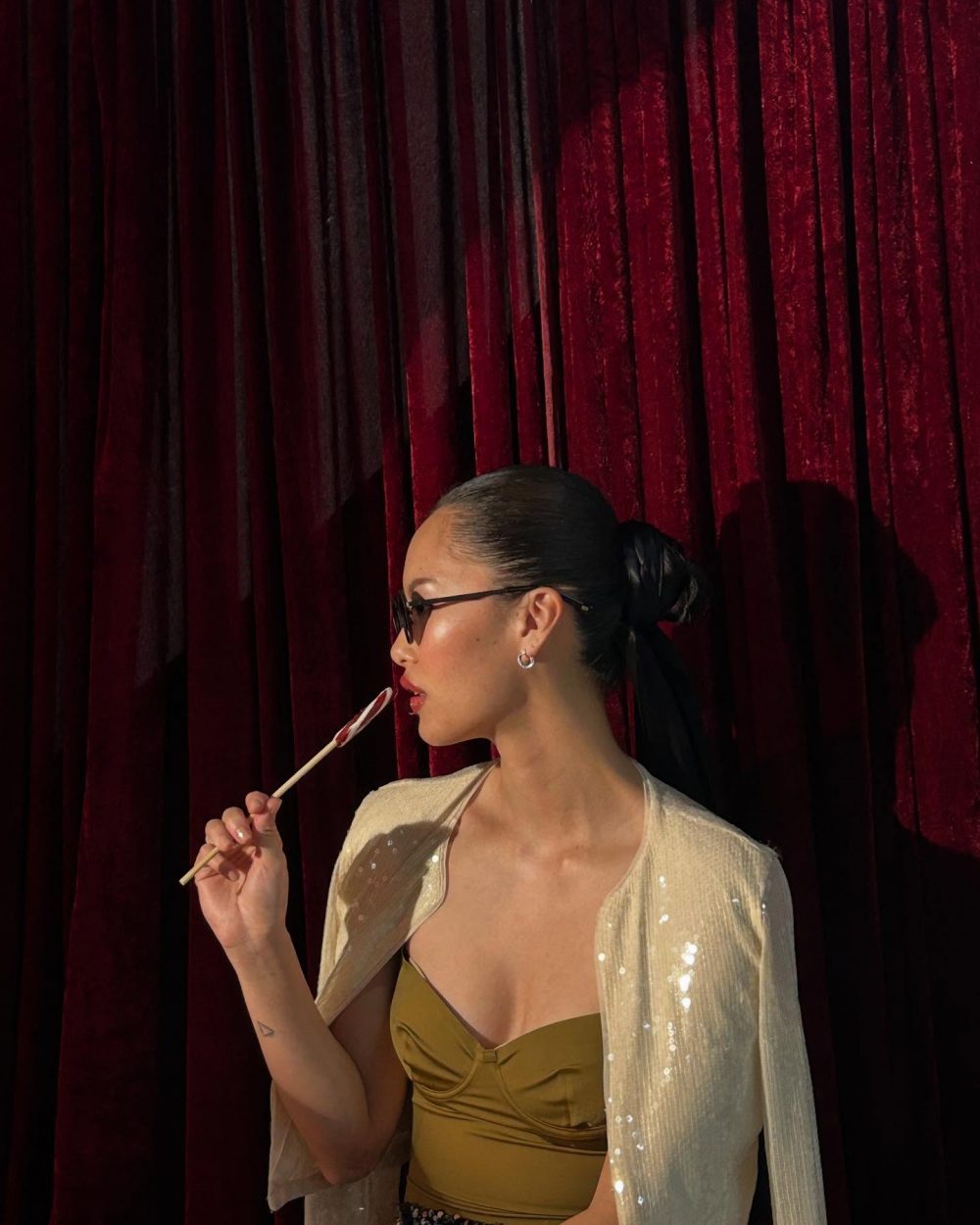 Woman in a gold dress with a sheer sequined shawl, sunglasses, and a lollipop against red velvet curtains.