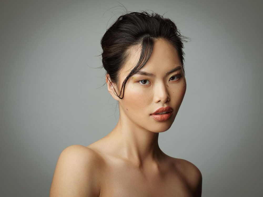 Portrait of an Asian woman with dark hair in a sleek updo, wearing subtle makeup and gold accents under the eyes, against a neutral gray background, bare shoulders visible.