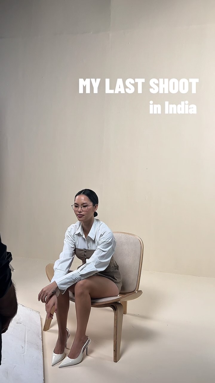 A woman in a white shirt and beige dress sits on a wooden chair in a studio; wall behind reads 'MY LAST SHOOT in India' in bold white letters.