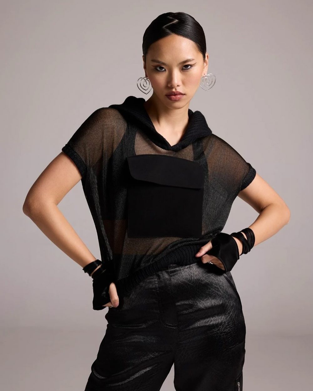 Model wearing a sheer black top with a large front pocket and loose black pants, accessorized with oversized heart-shaped hoop earrings, posing with hands on hips.
