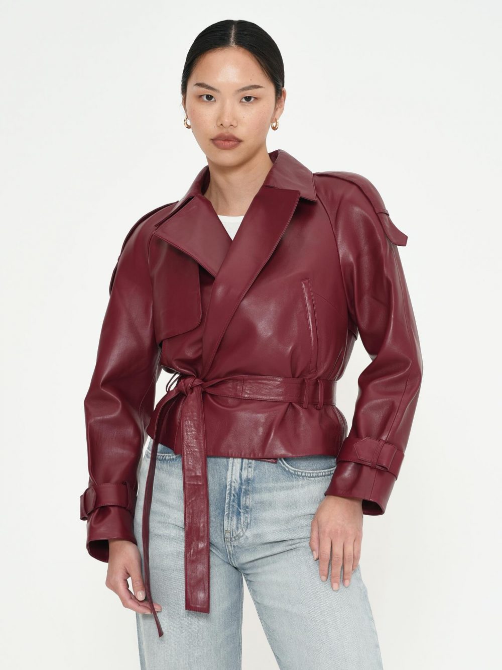 Woman in a burgundy leather belted trench jacket and light-wash jeans against a white background.