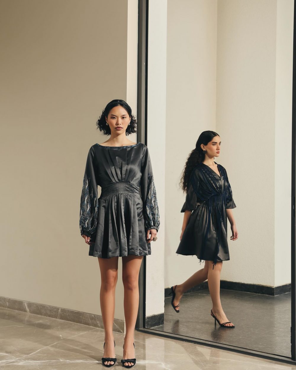 Two models in black satin dresses pose indoors beside a glass doorway, one facing the camera and the other in profile with heels.