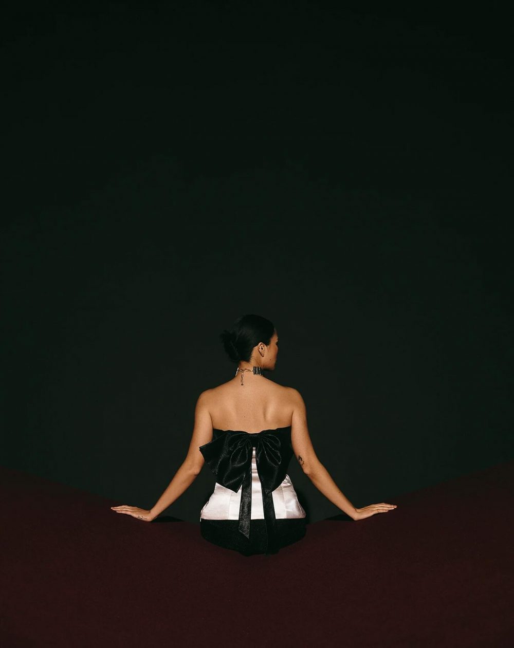 Woman seated with back to the camera on a dark stage, wearing a strapless white dress with a large black bow at the back.