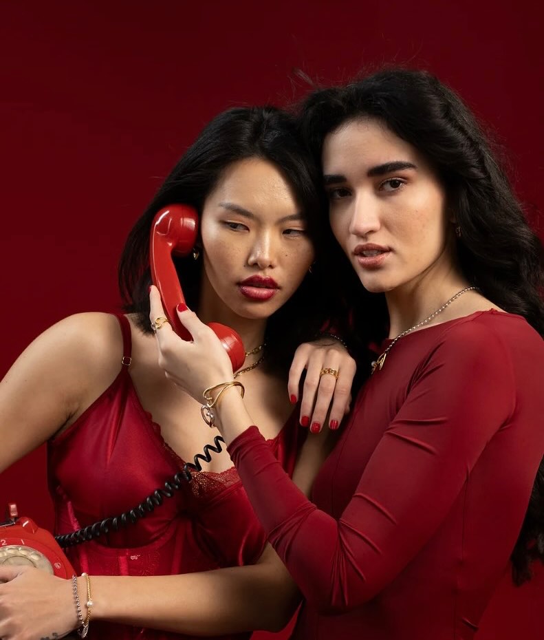 Two women in red dresses posing with a red rotary phone against a solid red backdrop.